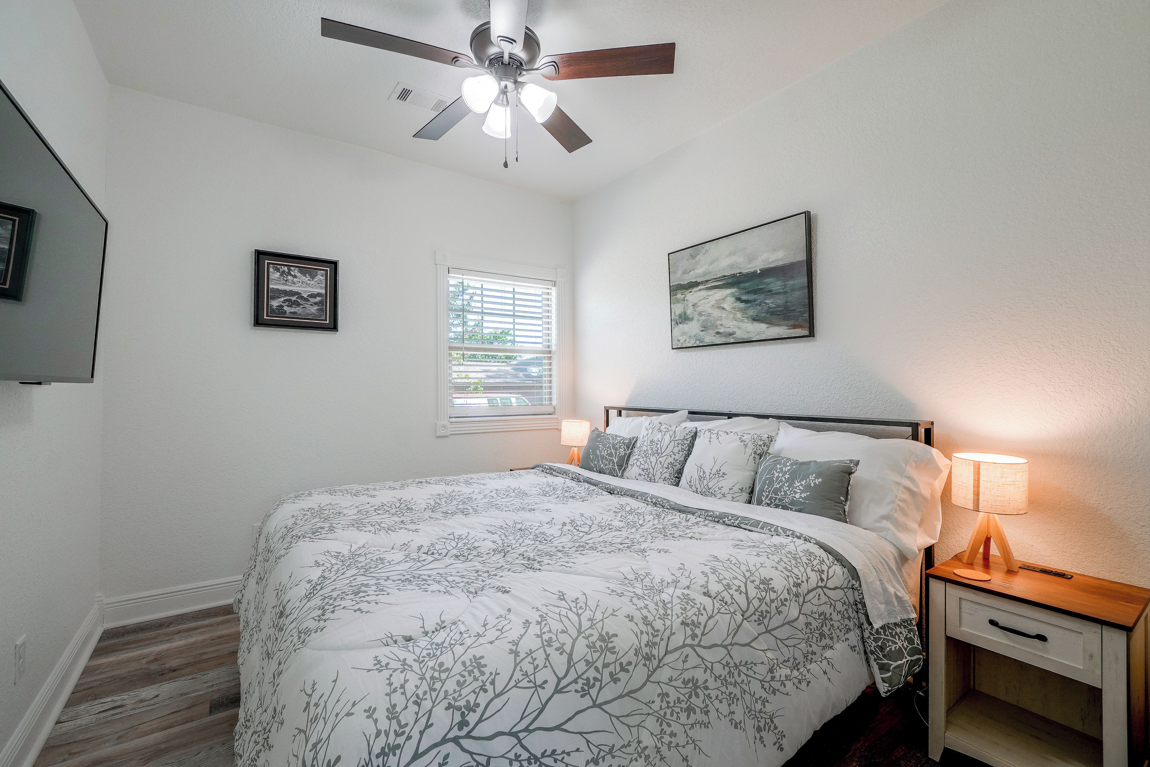 Une chambre à coucher avec un lit, un ventilateur de plafond, une fenêtre avec des stores, un tableau au mur et une lampe de chevet.