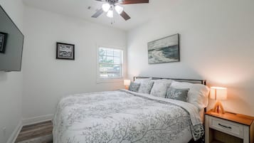 A bedroom with a bed, a ceiling fan, a window with blinds, a painting on the wall, and a bedside lamp.