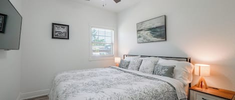A bedroom with a bed, a ceiling fan, a window with blinds, a painting on the wall, and a bedside lamp.