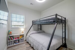 A bunk bed with a white mattress and grey bedding, a white shelving unit with books and toys, and a window with blinds. - Little Rock Family Home, 1 Mi to Dtwn (Little Rock)