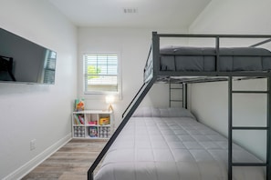 A bunk bed room with a white wall, a window with blinds, a small desk with a lamp, and a shelf with books and toys. - Little Rock Family Home, 1 Mi to Dtwn (Little Rock)