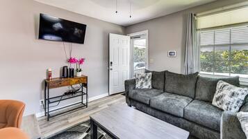 A living room with a grey sofa, a wooden coffee table, a TV mounted on the wall, and a window with blinds.