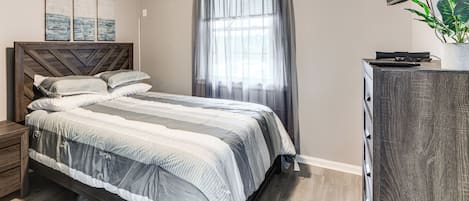 Un dormitorio con cama, mesita de noche, cómoda, televisor y ventana con cortinas.