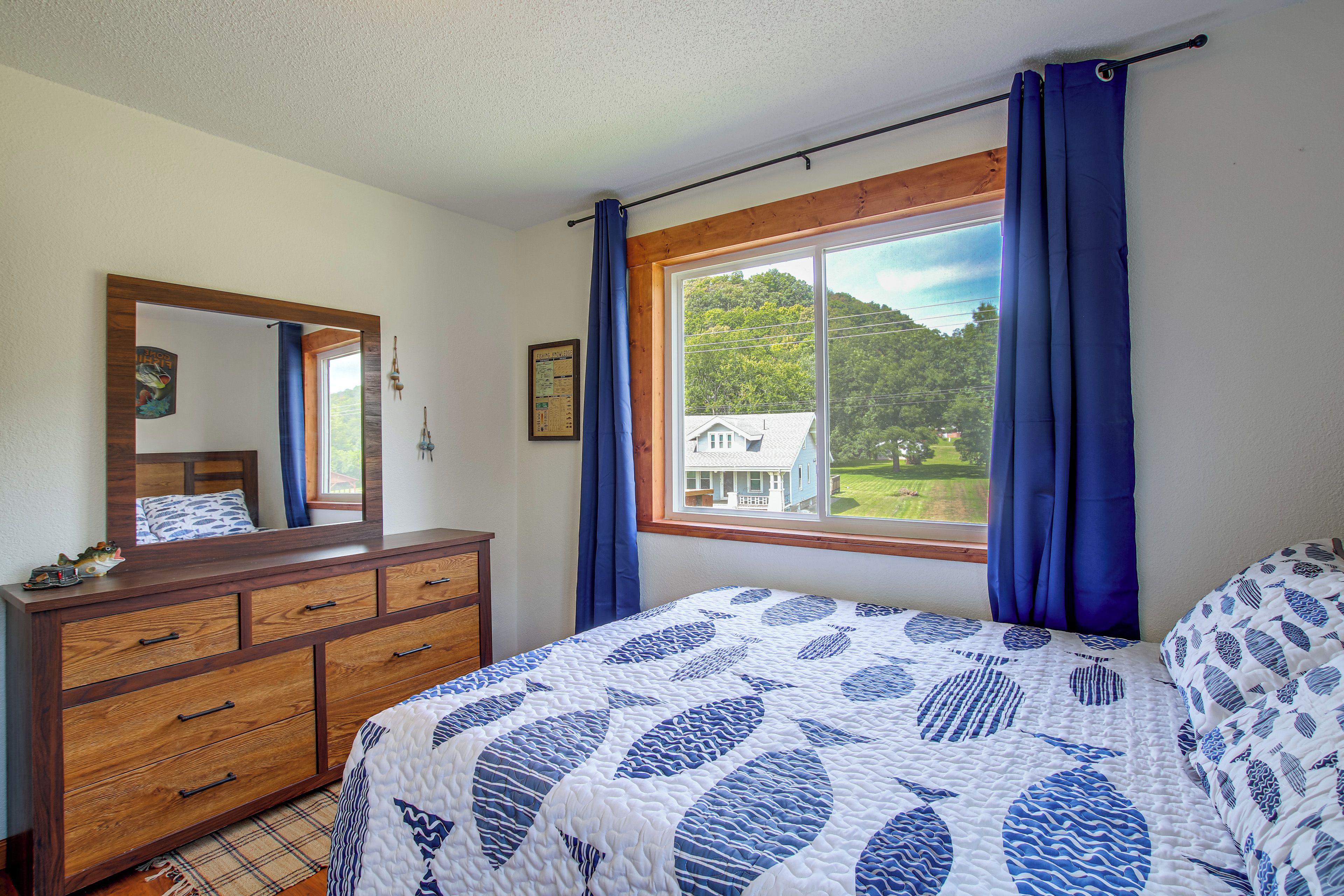 A bedroom with a wooden dresser, a bed with a patterned quilt, a window with curtains, and a view of a house and trees.