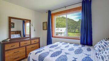 A bedroom with a wooden dresser, a bed with a patterned quilt, a window with curtains, and a view of a house and trees.
