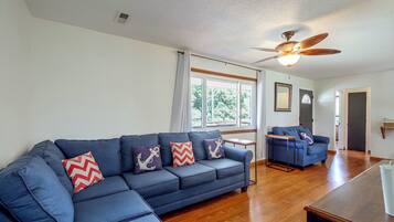 A living room with a blue sofa, wooden flooring, a ceiling fan, and a view through the windows.