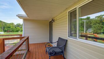 A covered porch with a wooden deck, a metal railing, a single chair, and a large window offering a view of a green landscape.