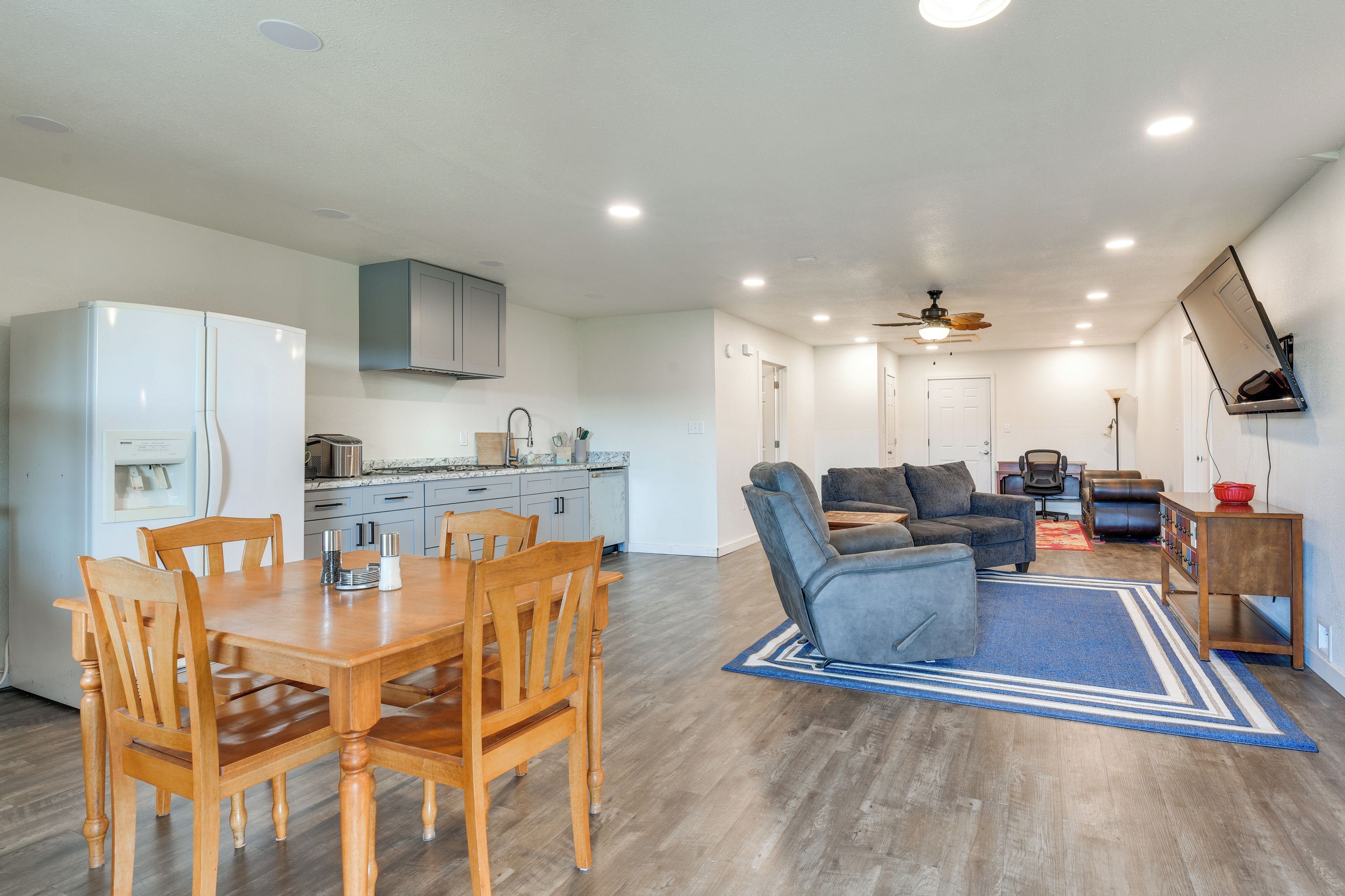A modern kitchen with built-in appliances, a dining area with wooden furniture, a gray sofa, and a ceiling fan.