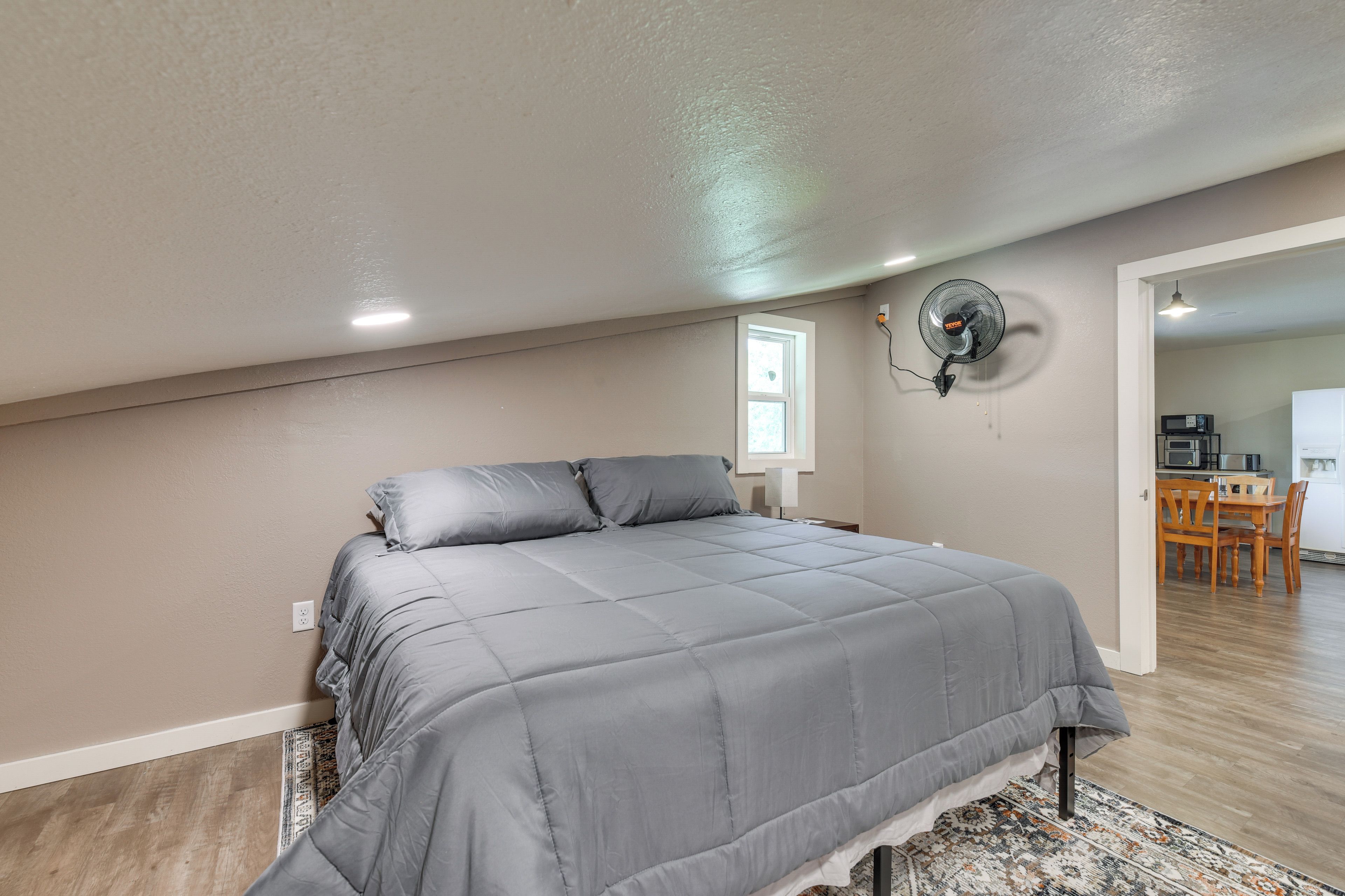 A bedroom with a bed, a wall-mounted fan, a window, and a view of a kitchen area.