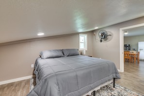 A bedroom with a bed, a wall-mounted fan, a window, and a view of a kitchen area.