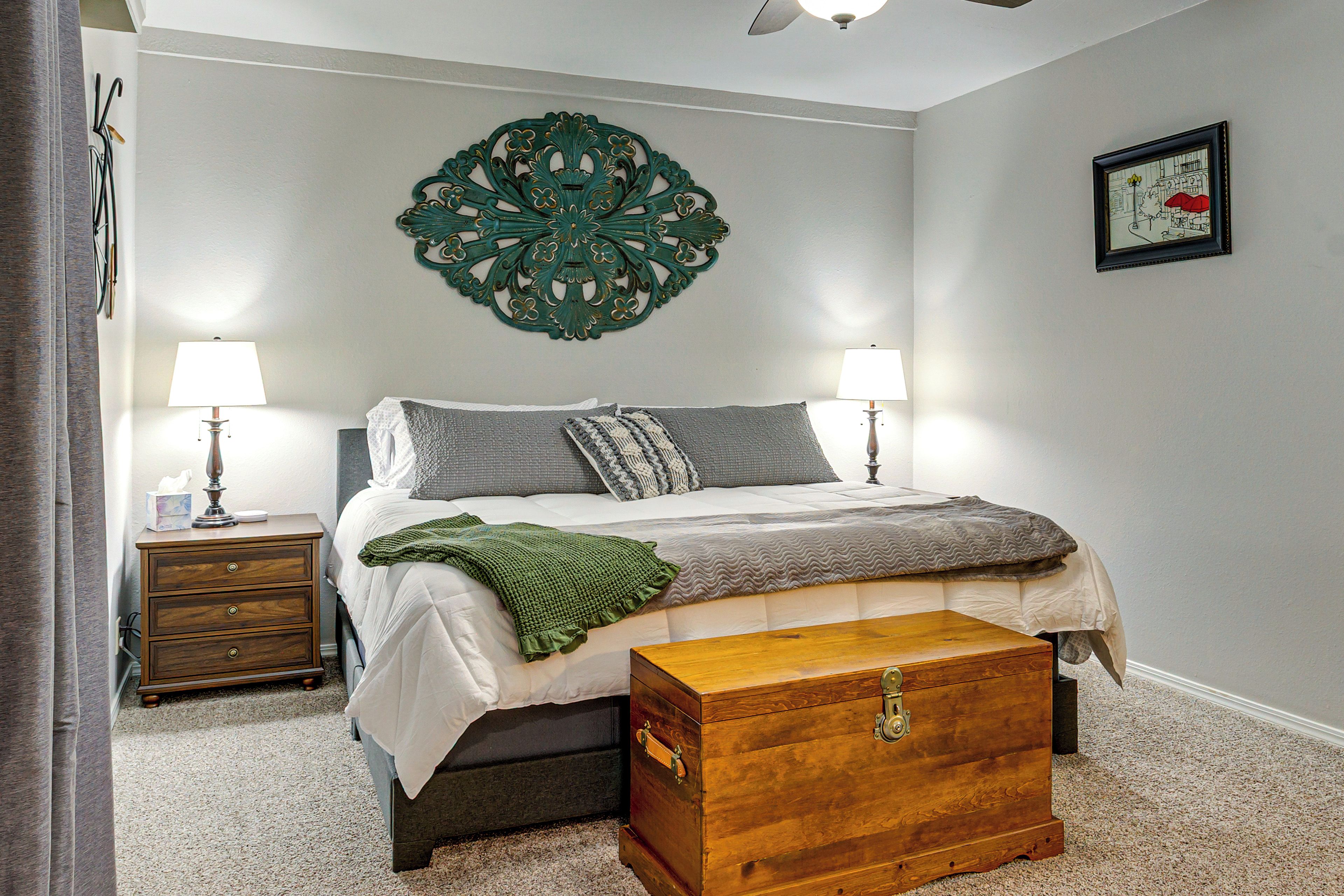 A bedroom with a bed, bedside lamps, a decorative wall piece, a framed picture, and a wooden chest.