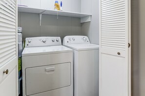 Une buanderie équipée de deux machines à laver et deux sèche-linge superposés, une étagère au-dessus pour ranger les produits de nettoyage, et une porte dotée d’une poignée en laiton.
