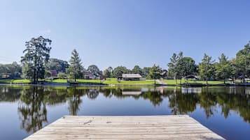A wooden dock extending into a calm lake with a clear reflection, surrounded by trees and a grassy area with a building in the background.