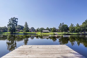 A wooden dock extending into a calm lake with a clear reflection, surrounded by trees and a grassy area with a building in the background. - Cast & Relax! Ventress River Home w/ Shared Pier (Ventress)