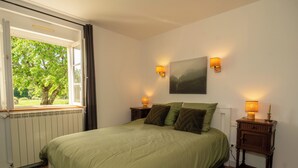 Quarto com cama, duas mesas de cabeceira, janela com cortinas, radiador e um quadro na parede.