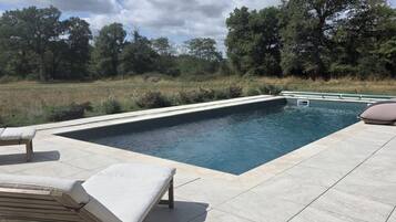 Une piscine rectangulaire, bordée d’une terrasse carrelée, entourée d’arbres et d’un champ d’herbe.