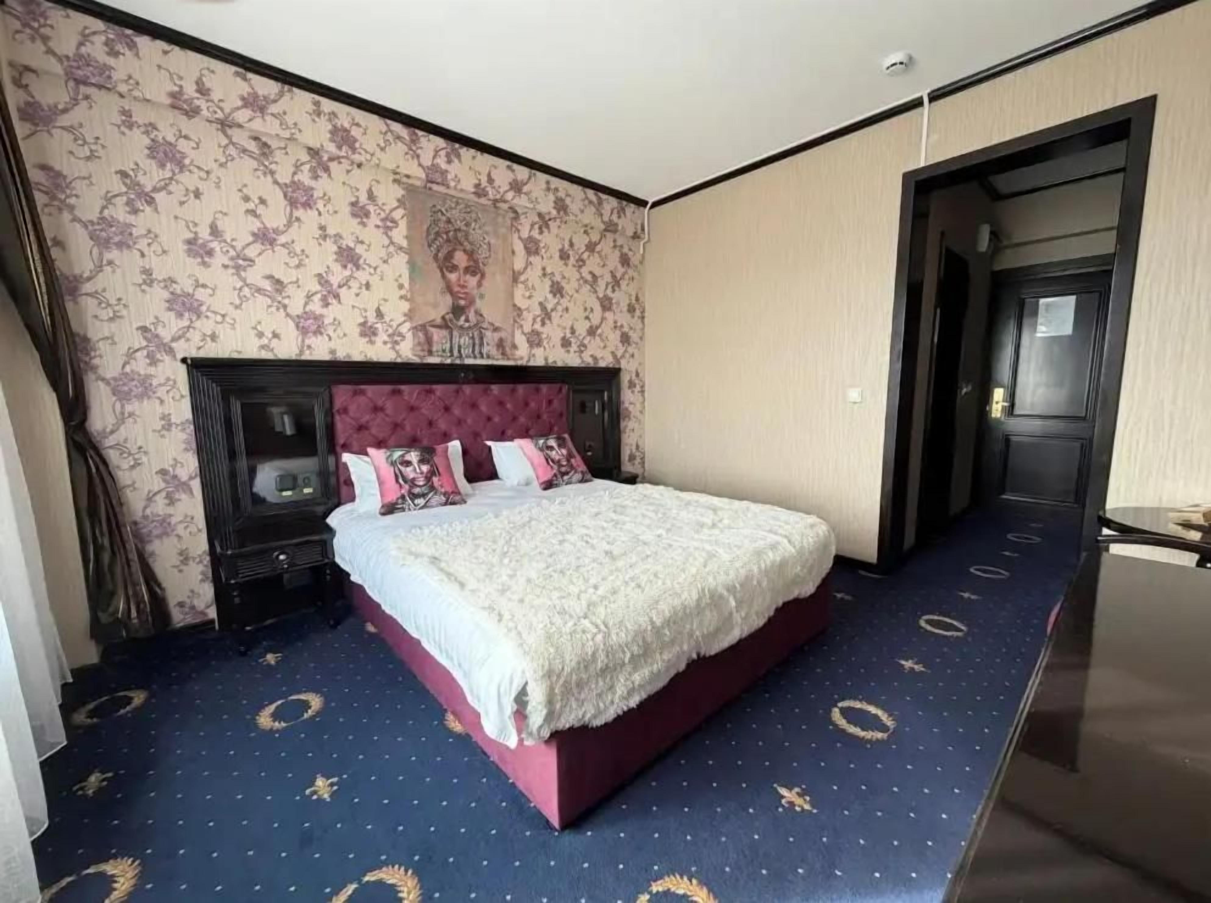 Quarto de hotel com uma cama grande, cabeceira escura, mesa de cabeceira e televisão na parede.