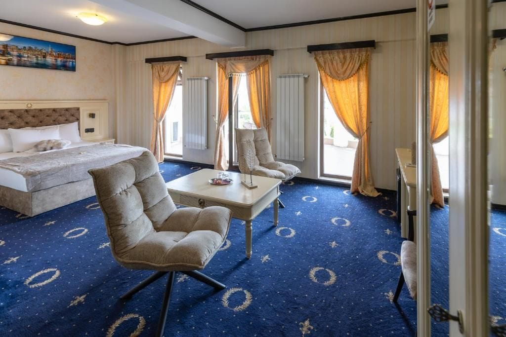 A hotel room with a large bed, a seating area with a table, and a view of the outdoors through the windows.