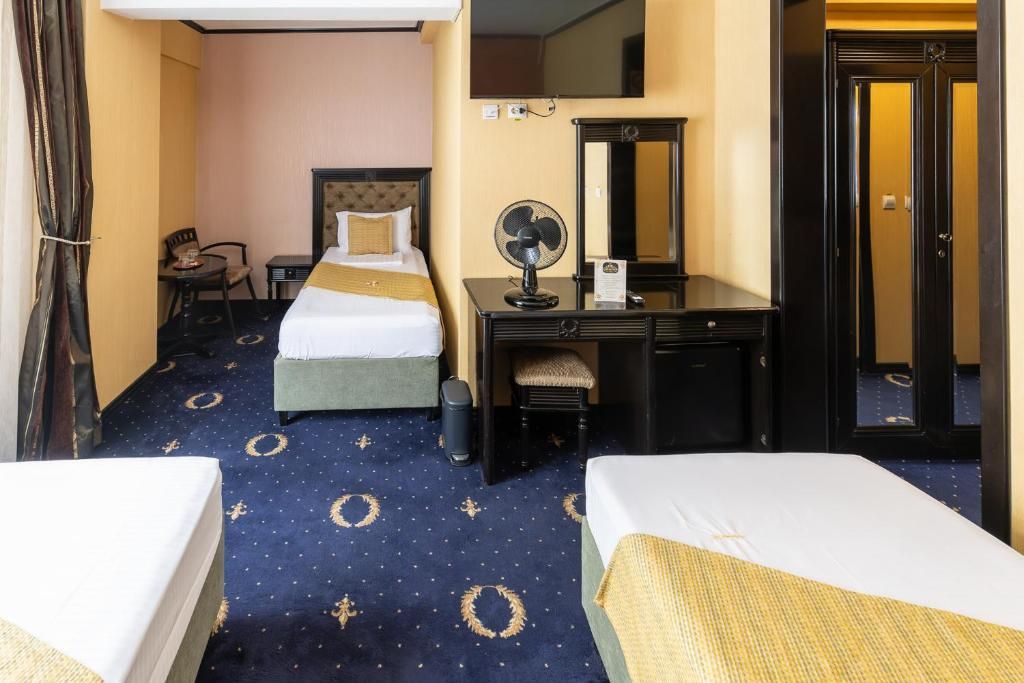 A hotel room with two beds, a desk with a chair, a small table, a mirror, a fan, and a door.