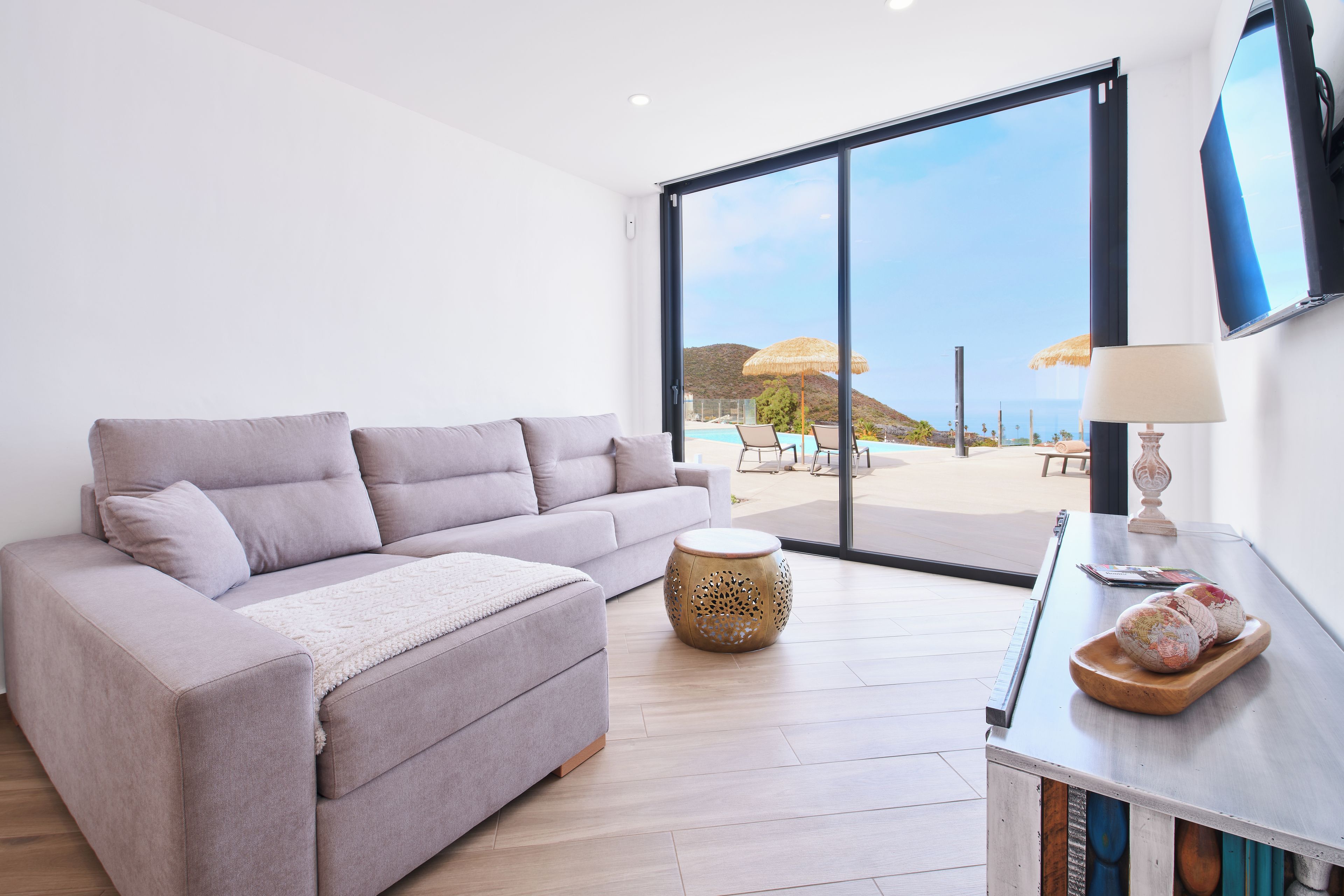 Un salón moderno con un sofá gris, una mesita redonda de madera y vistas a una piscina y una playa a través de puertas de cristal amplias.