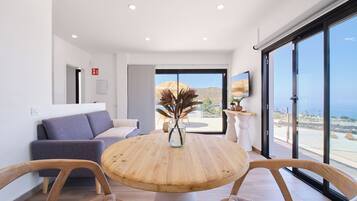A modern living room with a round wooden table, two wooden chairs, a grey sofa, and a large window offering a view of the sea.