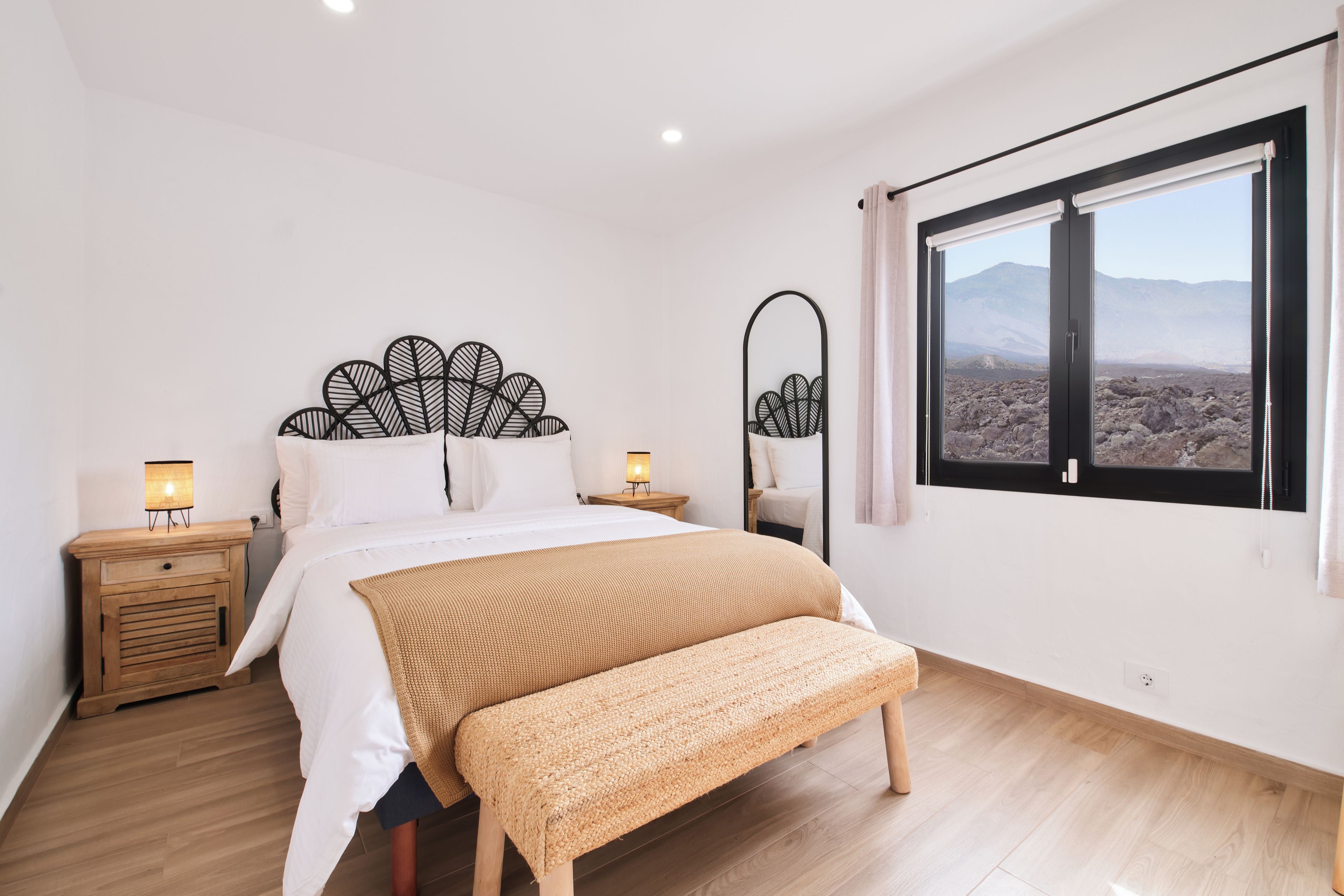Un dormitorio con cama, mesitas de noche, un banco, una ventana con vistas y un espejo montado en la pared.