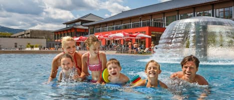 A swimming pool with people in the water, a building with red umbrellas in the background, and a water feature.