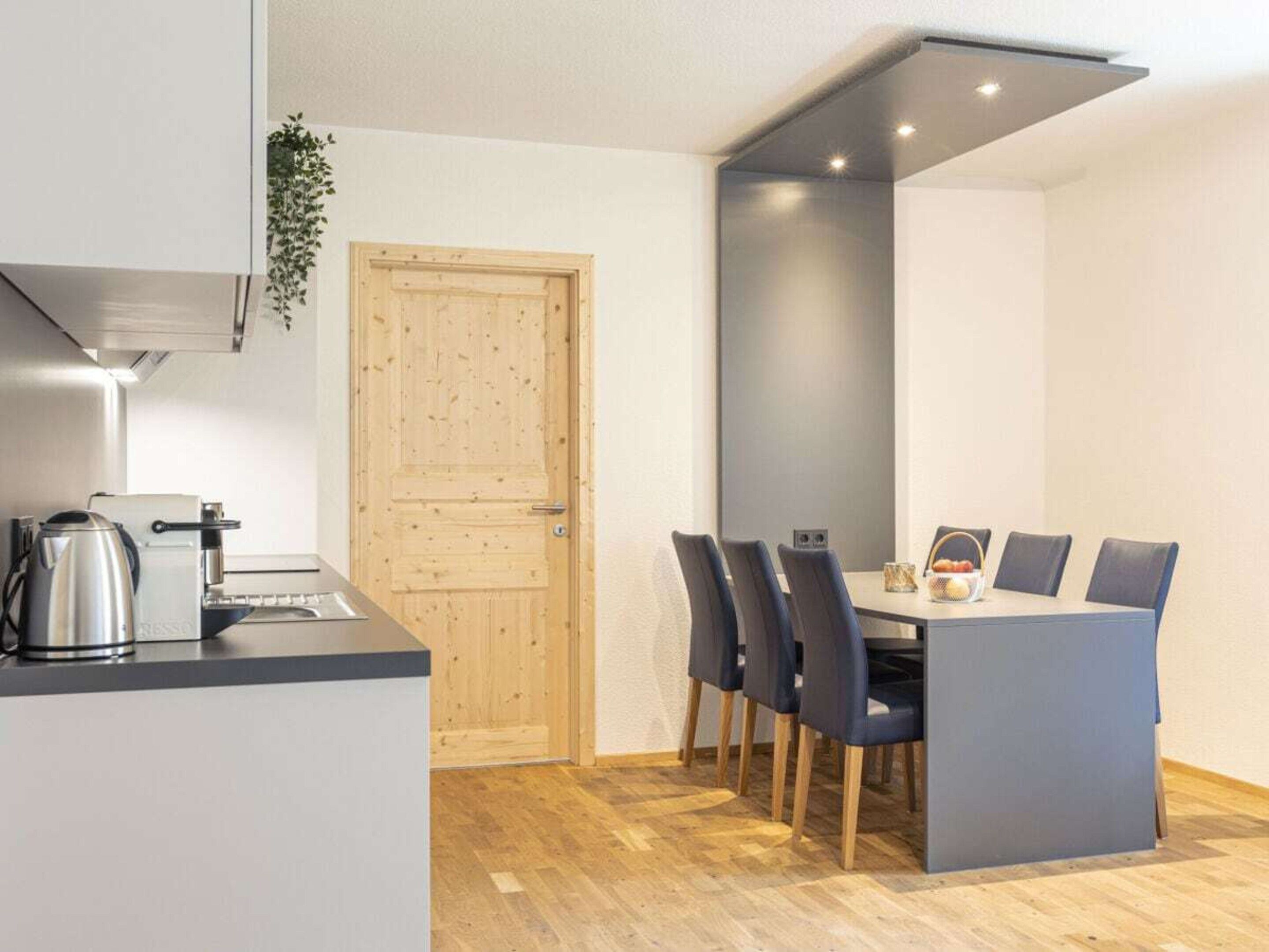A compact kitchen with a coffee maker, a wooden door, and a dining area with a table and chairs.