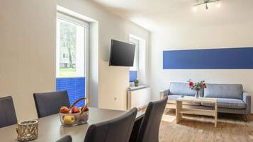 A modern living room with a dining area, a flat-screen TV mounted on the wall, and a sofa with a vase of flowers.