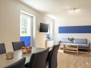 Un salon moderne comprenant un coin repas, un téléviseur à écran plat fixé au mur et un canapé agrémenté d’un vase de fleurs.
