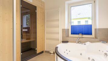 Une salle de bain avec une grande baignoire, une fenêtre avec des stores et une porte en bois donnant sur une autre pièce.