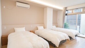 Habitación de hotel con tres camas individuales, aire acondicionado en el techo y un gran ventanal con persianas.