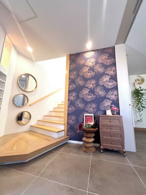 A staircase with wooden steps and a wall with a floral pattern. There are three round mirrors on the wall, a small wooden dresser, and a framed picture with flowers. - 30-second walk to the beach. Stay like you're living here. Stunning sunset and ocean views. Beachside Island Home, Steps from the Sea (Miyakojima)
