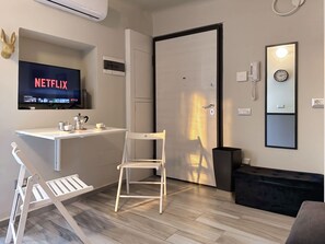 A modern hotel room with a flat-screen TV displaying Netflix, a desk with a coffee maker, a chair, a black sofa, and a mirror with a clock. - The 6th - Studio Apartment Porta Venezia (Milano)