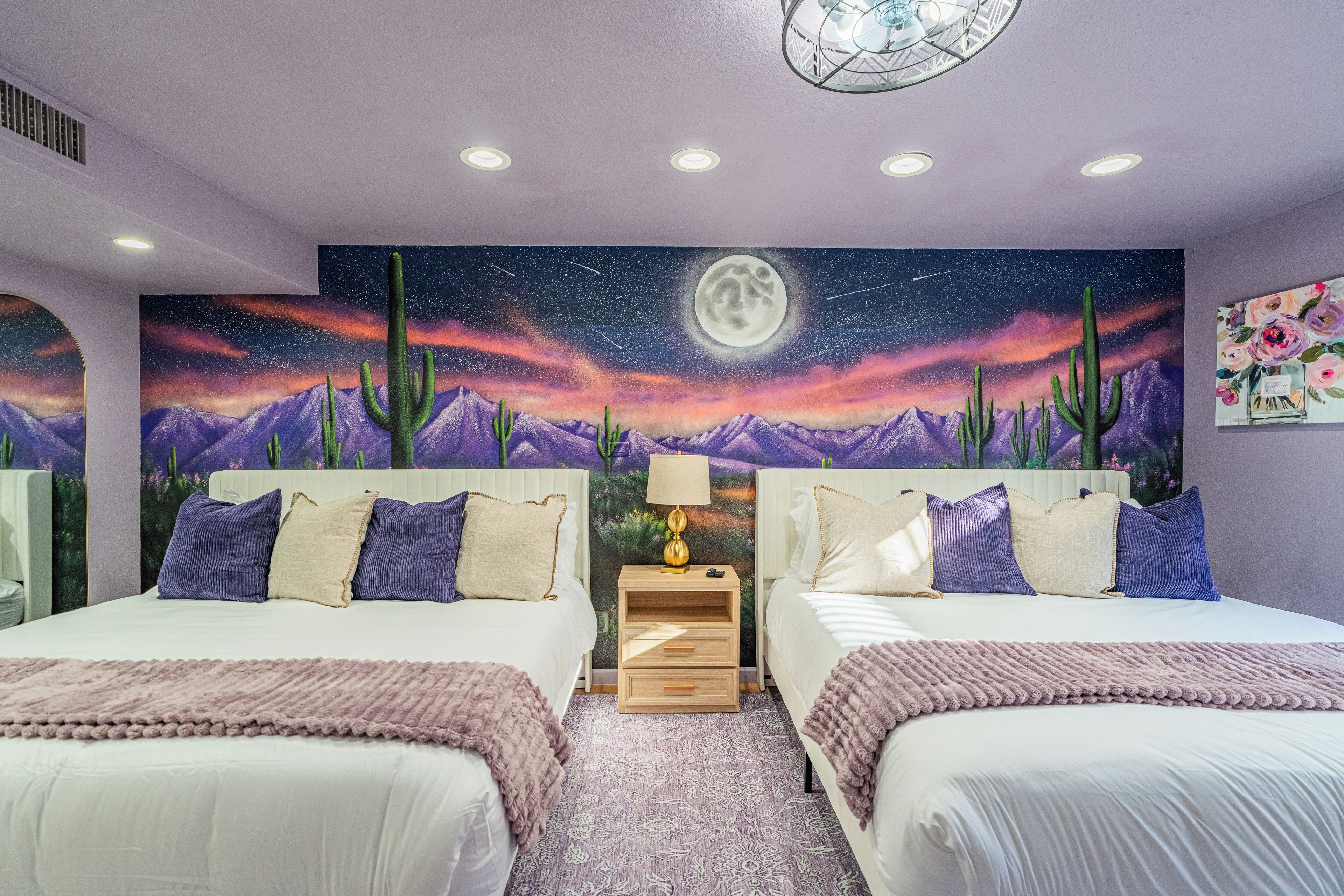 Een slaapkamer met twee bedden, een nachtkastje, een lamp en een muurschildering van een sterrenhemel met cactussen en een maan.