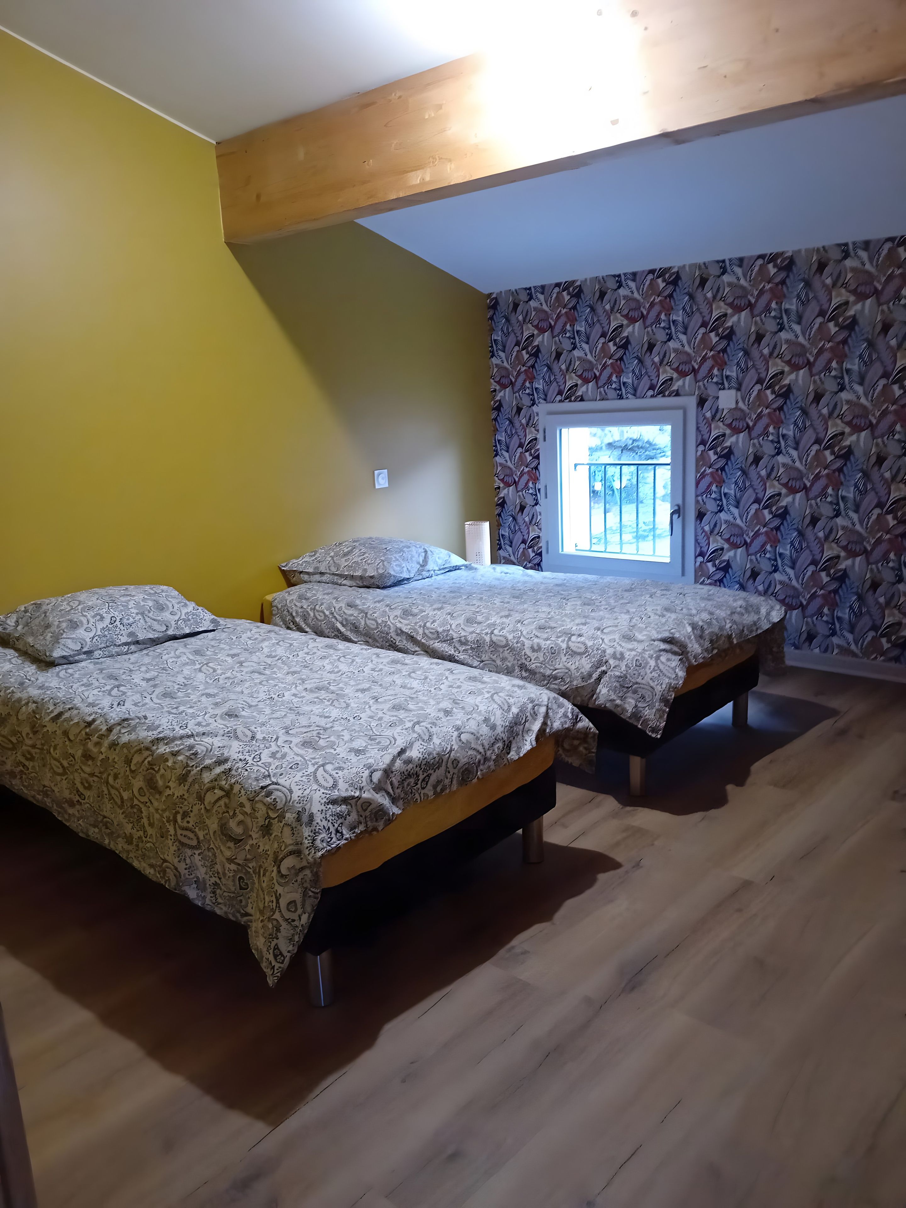 A room with two beds, wooden flooring, a window with a view, and a patterned wall.