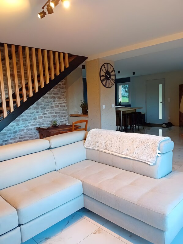 A modern living room with a sectional sofa, a wooden staircase, and a dining area in the background.