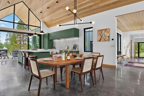 A modern kitchen with a central island, dining area with a round table and chairs, and large windows offering views of the outdoors. - Two Sisters Secludes private home mt views in-floor heating family rental (Tabernash)
