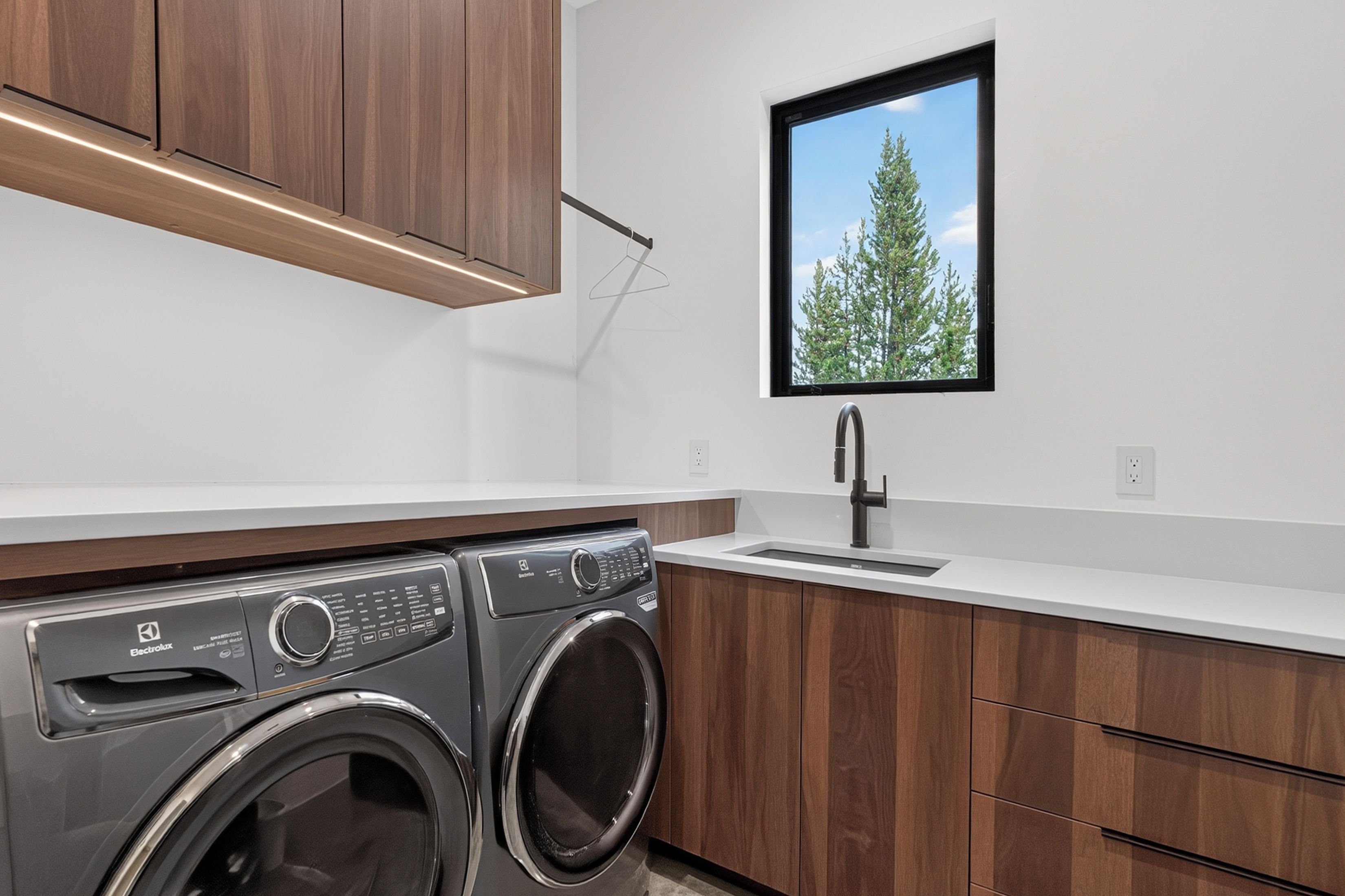 A modern laundry room with a front-loading washer and dryer, a sink, and a window with a view of trees.
