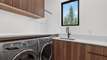 A modern laundry room with a front-loading washer and dryer, a sink, and a window with a view of trees.