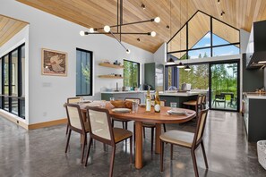 A modern kitchen with a dining area, large windows, and a wooden ceiling.