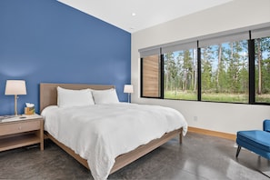 A bedroom with a large bed, two bedside tables, a nightstand, a blue chair, and a window with a forest view. - Two Sisters Secludes private home mt views in-floor heating family rental (Tabernash)