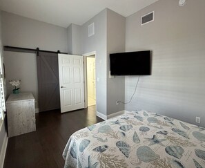 Quarto com cama, televisão fixada na parede, porta dando acesso a outro cômodo e uma pequena mesa com uma planta.