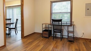 Ein Zimmer mit einem Holztisch, zwei Stühlen, einem Fenster mit Jalousien und einer Tür zu einem anderen Raum.