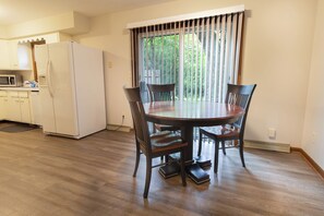 Un comedor con una mesa redonda y sillas, una cocina con refrigerador y microondas, y un ventanal con persianas.
