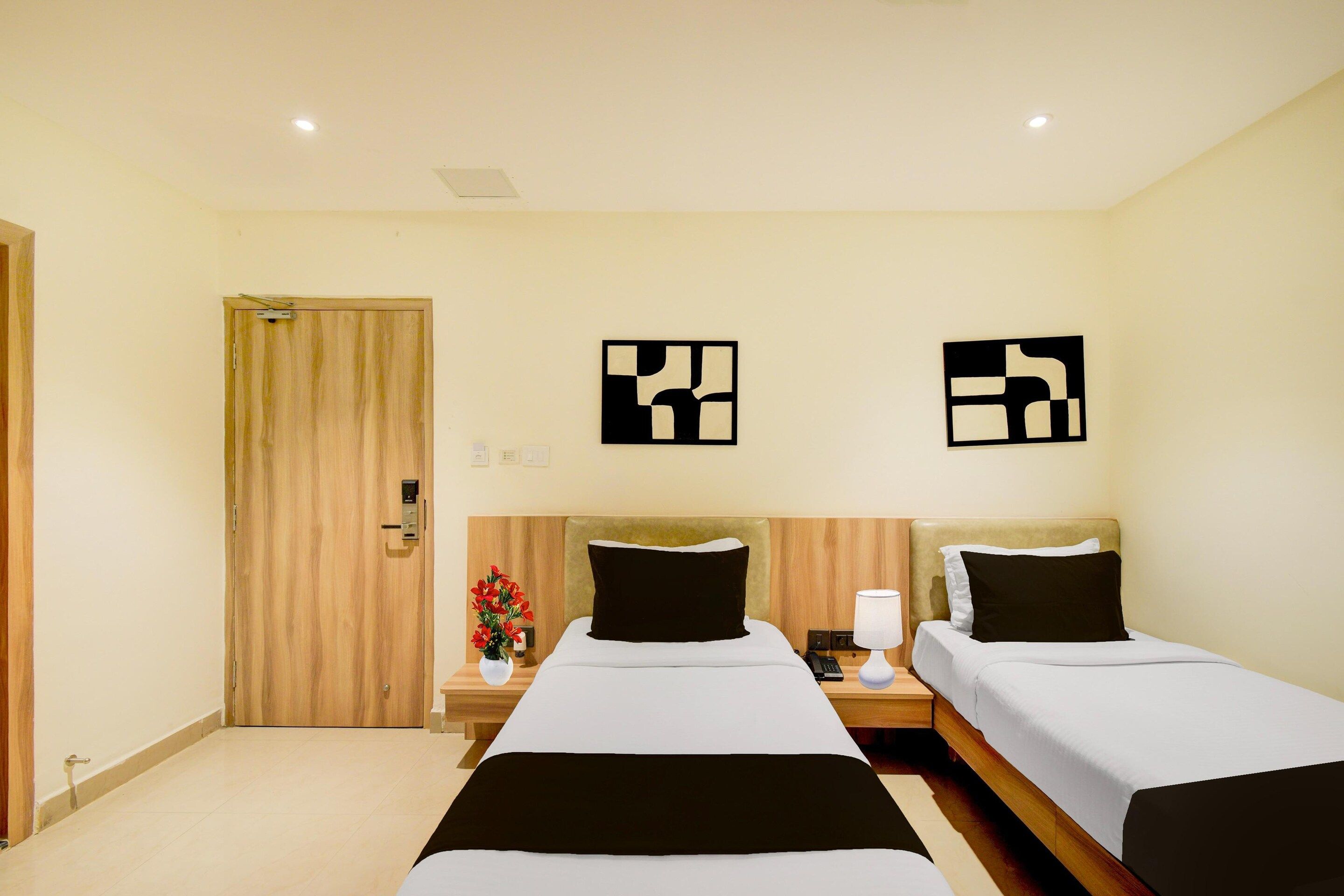A hotel room with two beds, a wooden door, a nightstand with a lamp, and two wall art pieces.