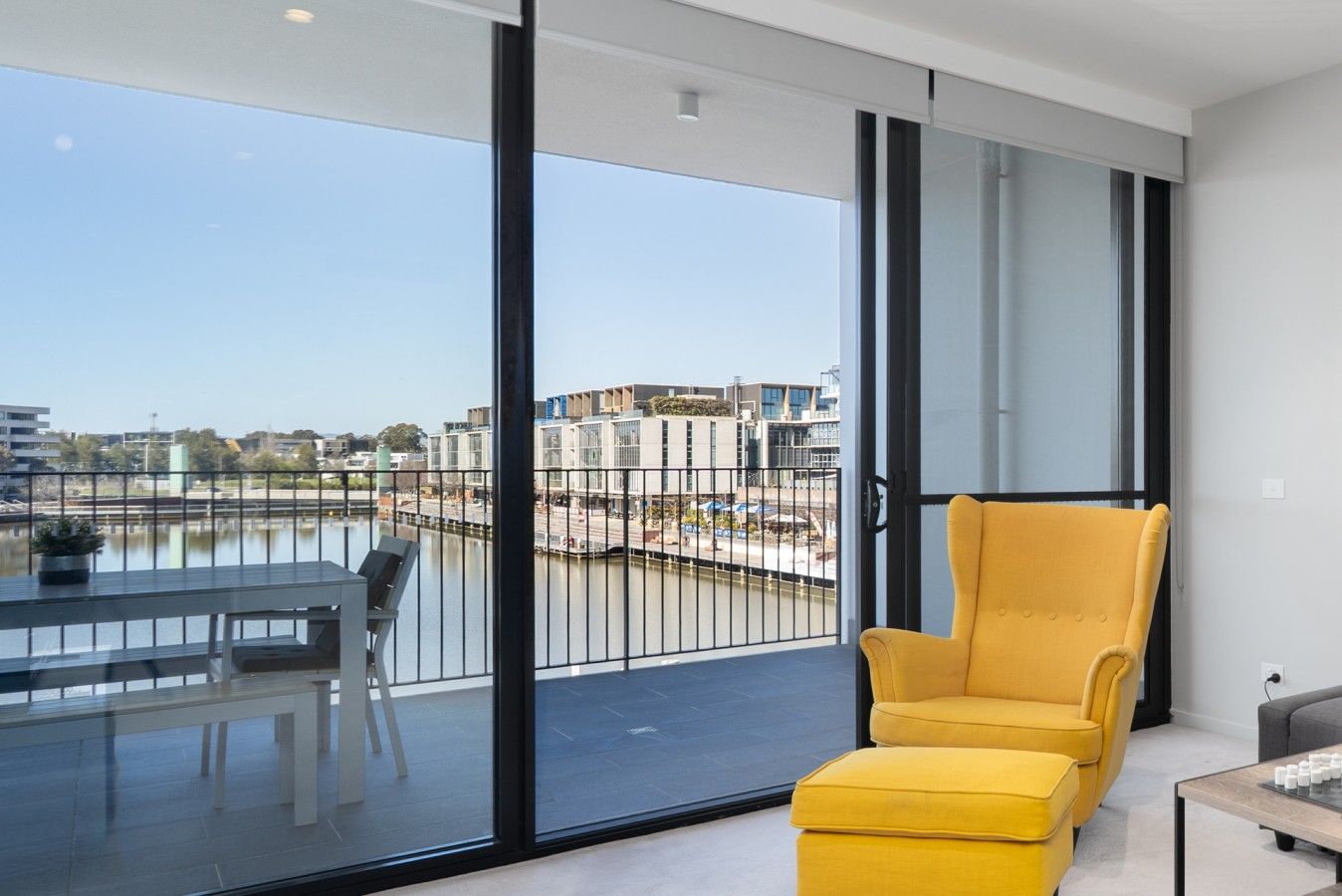 Un interior moderno con un sillón amarillo, una mesa de comedor con sillas y vistas a un río y edificios a través de puertas de cristal grandes.