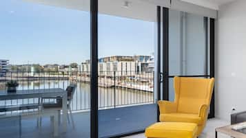 Un interior moderno con un sillón amarillo, una mesa de comedor con sillas y vistas a un río y edificios a través de puertas de cristal grandes.