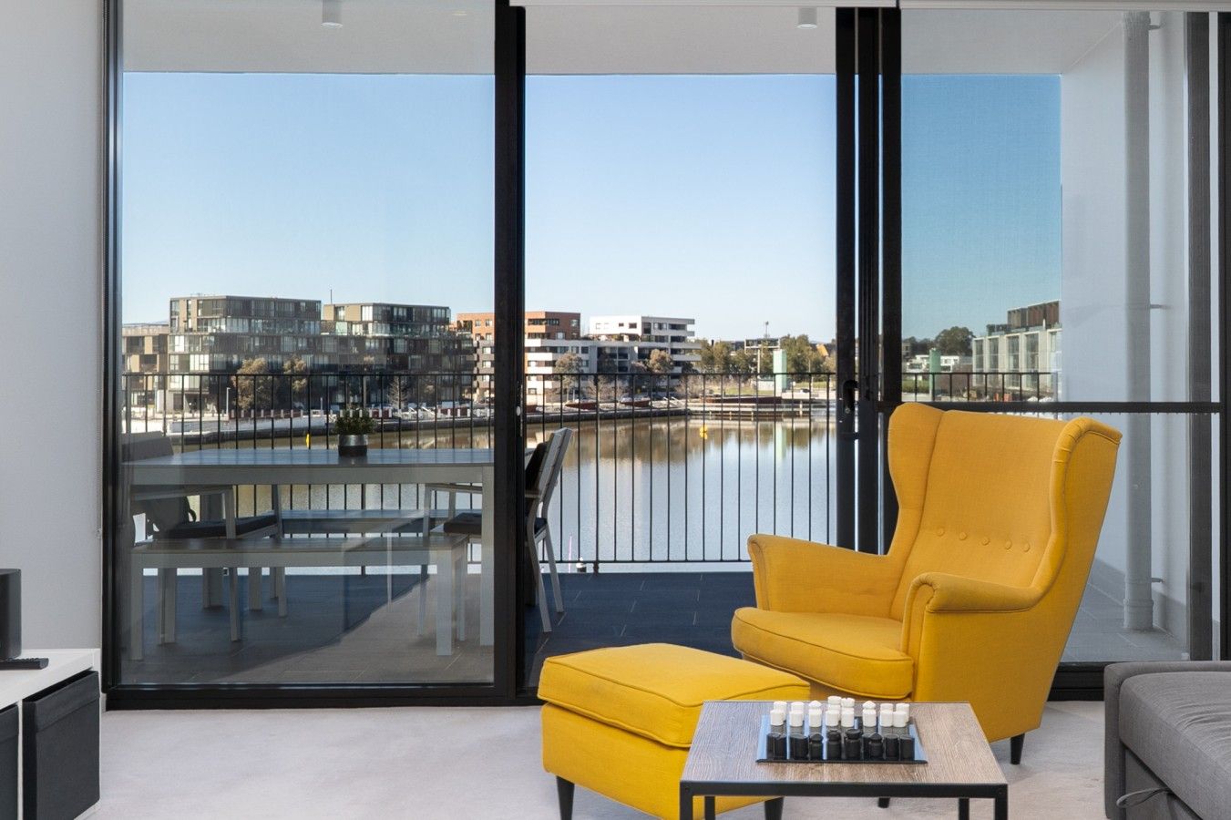 A modern balcony with a yellow armchair, a small table, and a view of a cityscape and water body.
