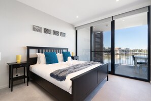 Un dormitorio moderno con una cama grande, una mesita de noche y vistas a un paisaje urbano desde un balcón.
